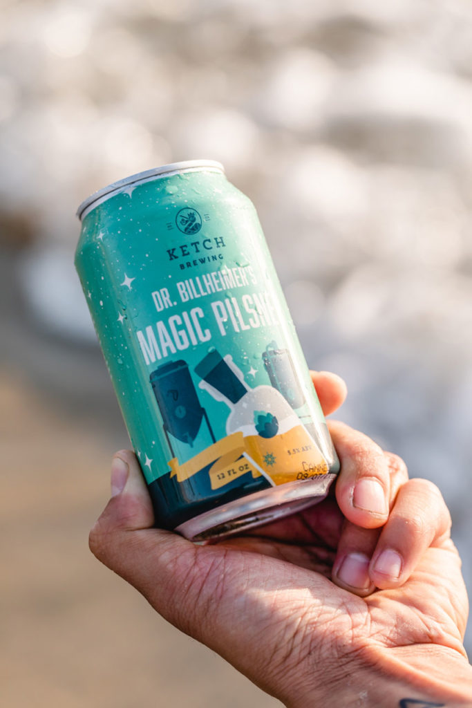 hand holding can of ketch magic pilsner with beach in background