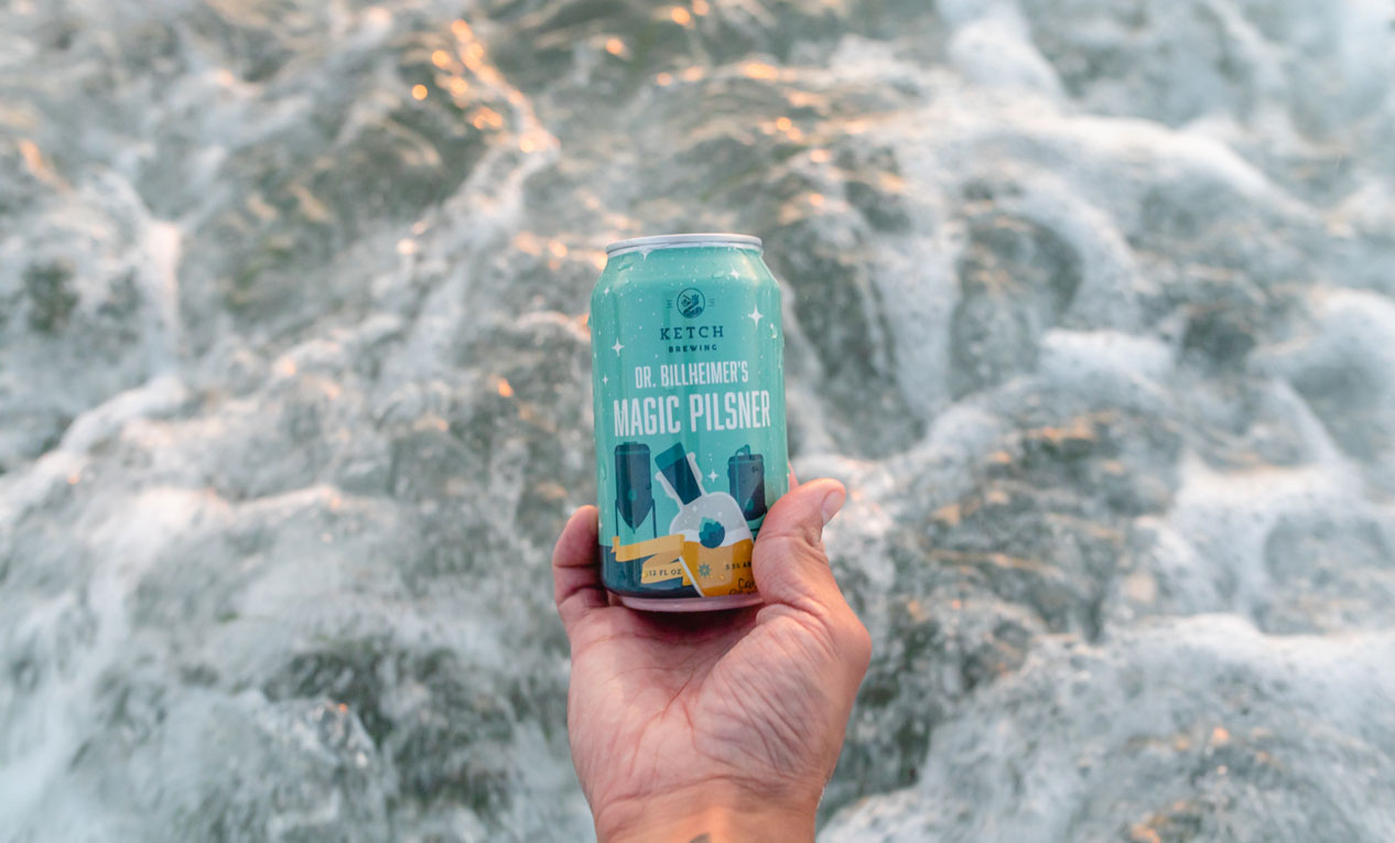 hand holding can of ketch magic pilsner in front of ocean waves