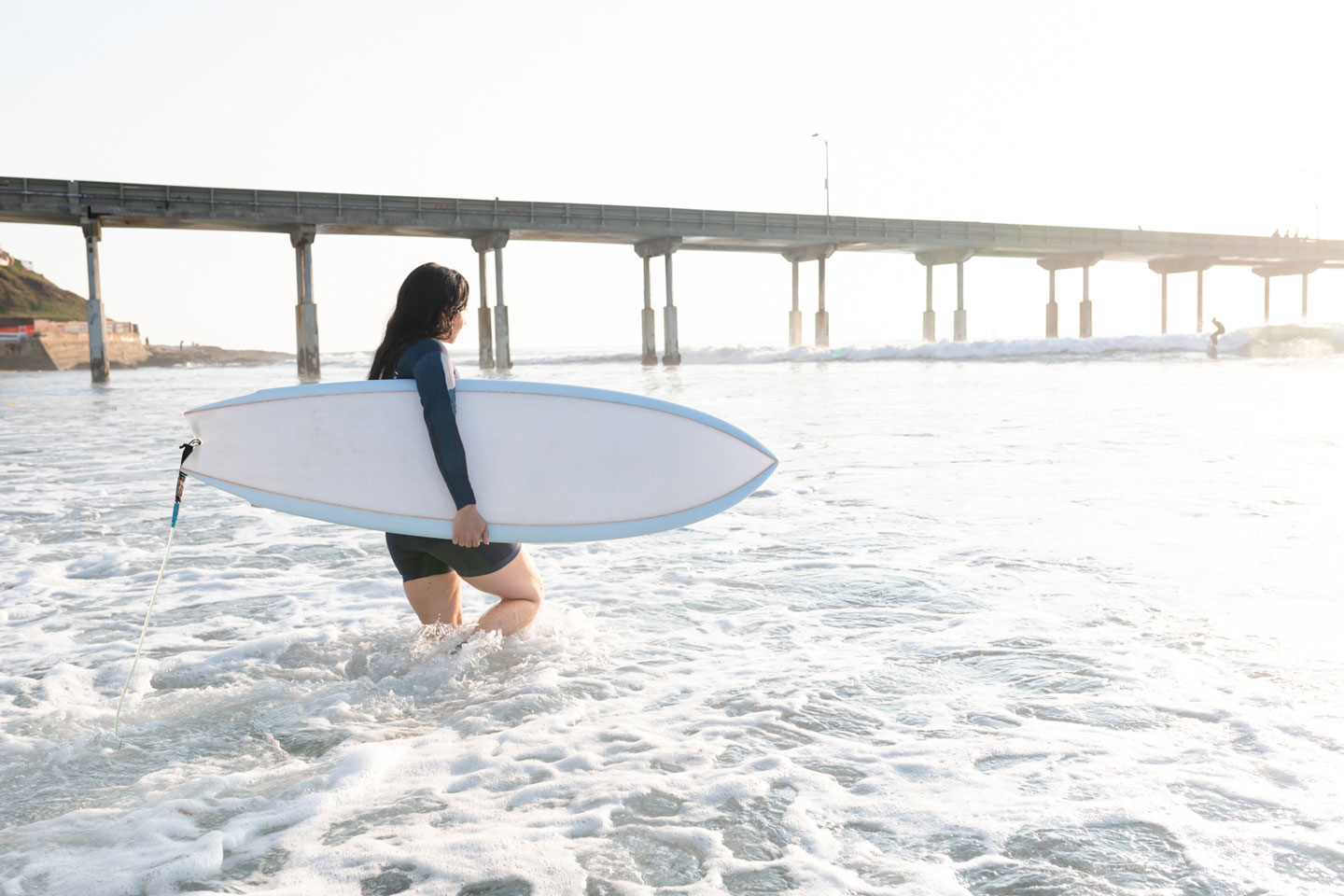 female surfer wading out into waves with surfboard under arm, with pier in the background